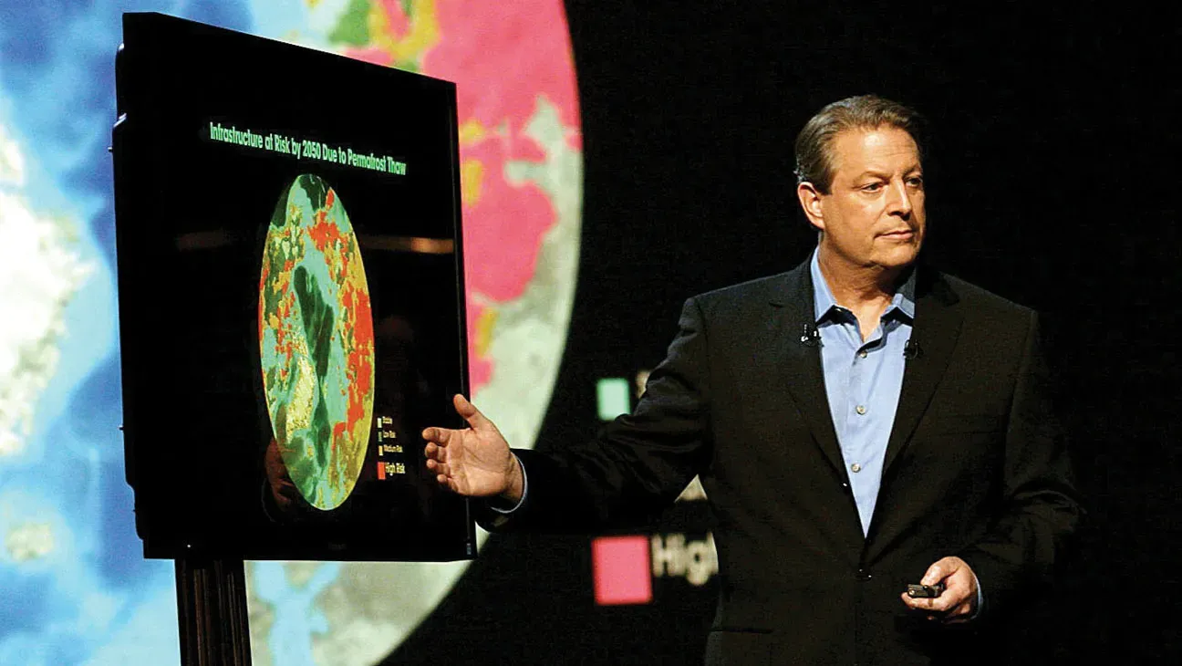 A picture of Al Gore standing in front of and gesturing to a TV screen that shows a color-coded version of Earth on it.