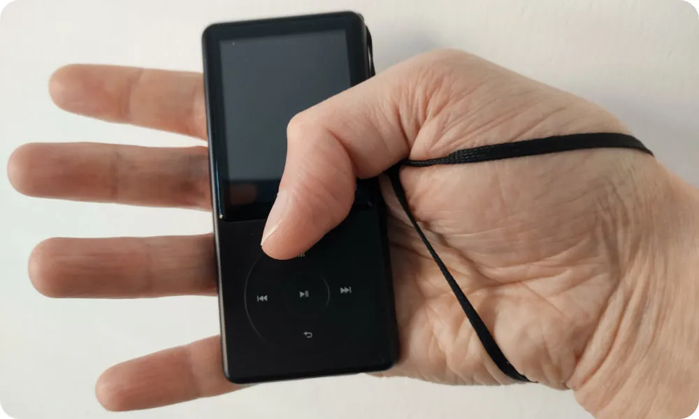 The MP3 player is attached via the strap to a person’s hand, the person’s thumb barely reaches the play button.