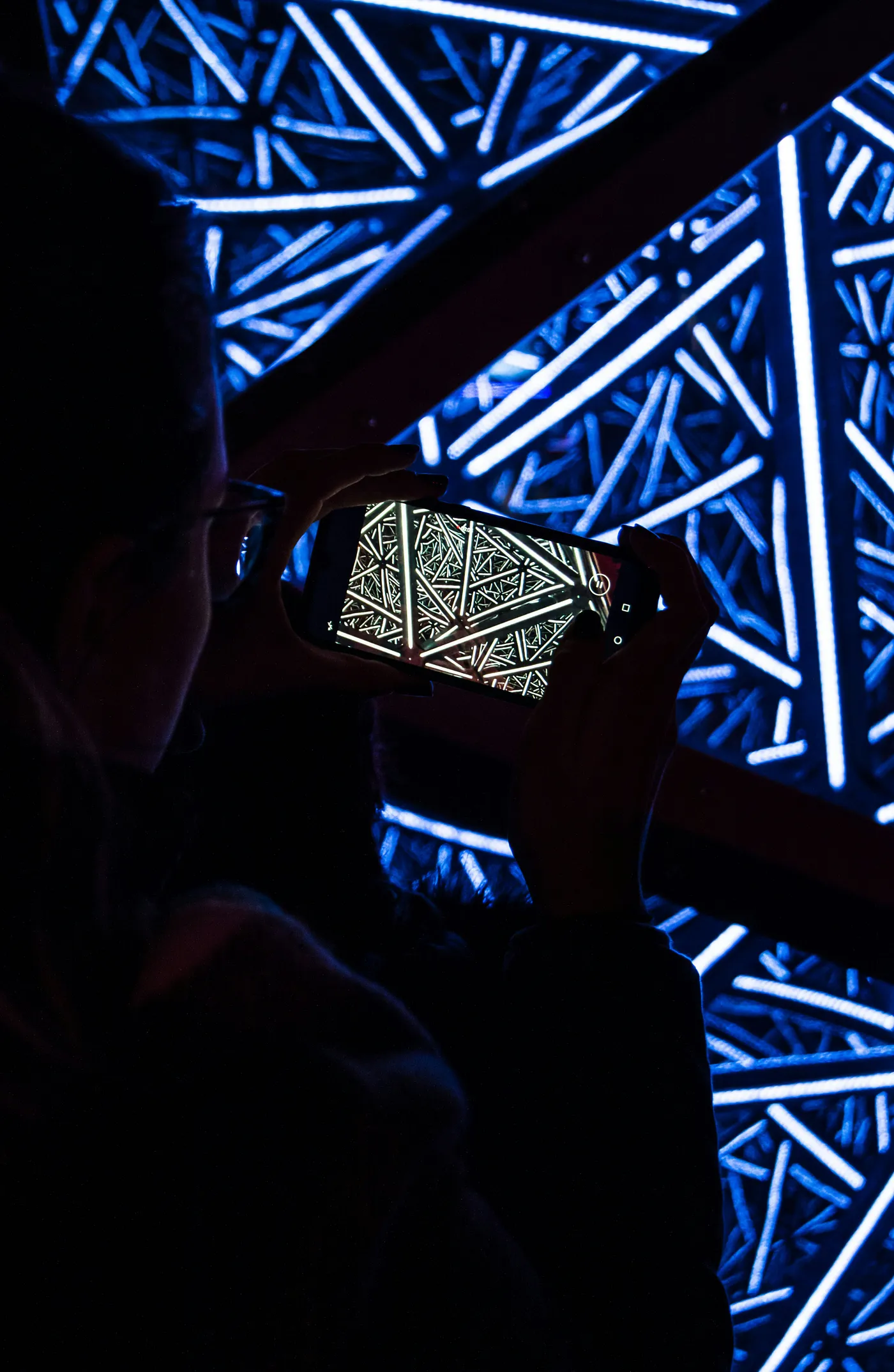 Person taking a photo of a bright blue light installation with intricate geometric patterns, reflected on the smartphone screen.