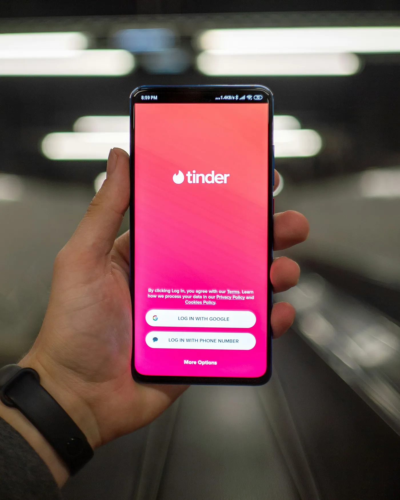 A hand holding a smartphone displaying the Tinder login screen, with options to log in via Google or phone number, against a blurred background