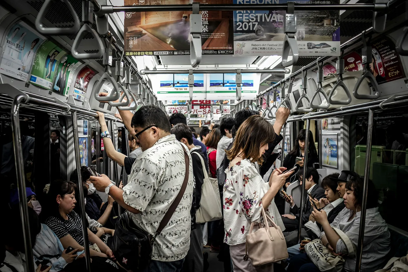 A crowded subway car with passengers standing and sitting, most of them looking at their smartphones, surrounded by advertisements and handrails.