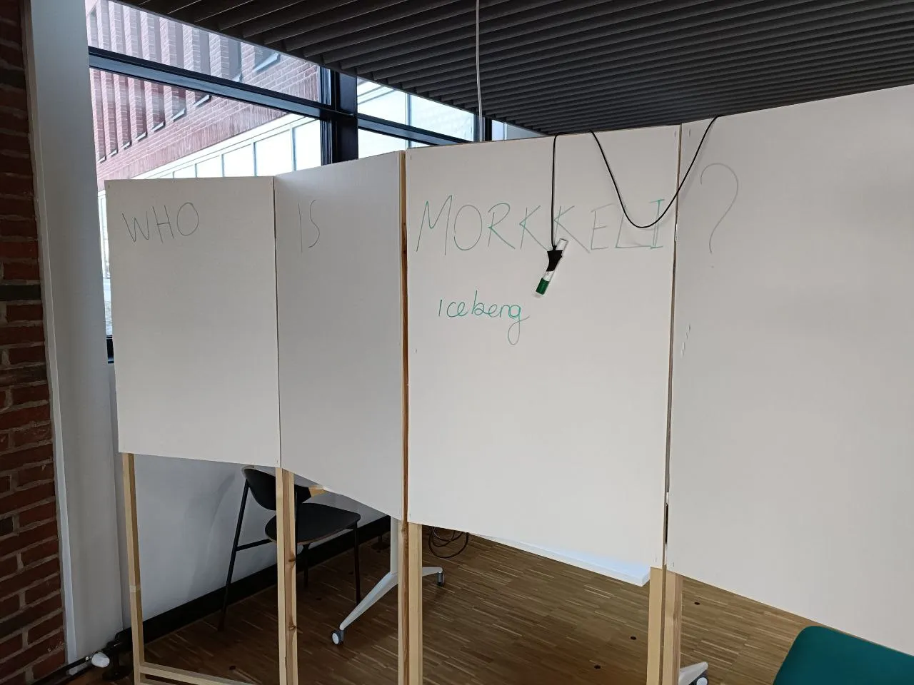 A folding screen with a haphazardly attached marker on it. The screen has a large text “WHO IS MORKKELI?” written on it. Underneath it, there is a smaller text: “Iceberg.”